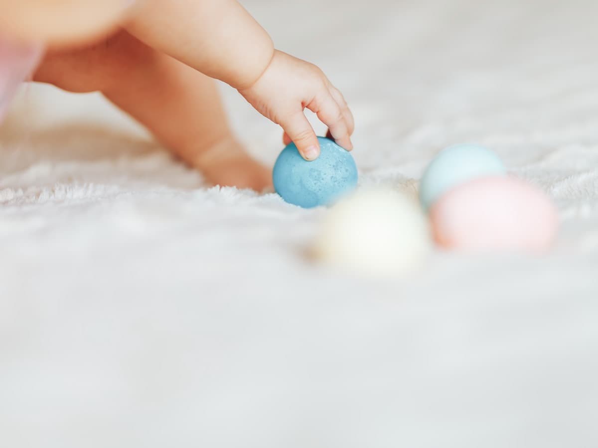 Eine Babyhand greift nach pastellfarbenen Eiern auf einer weichen Decke